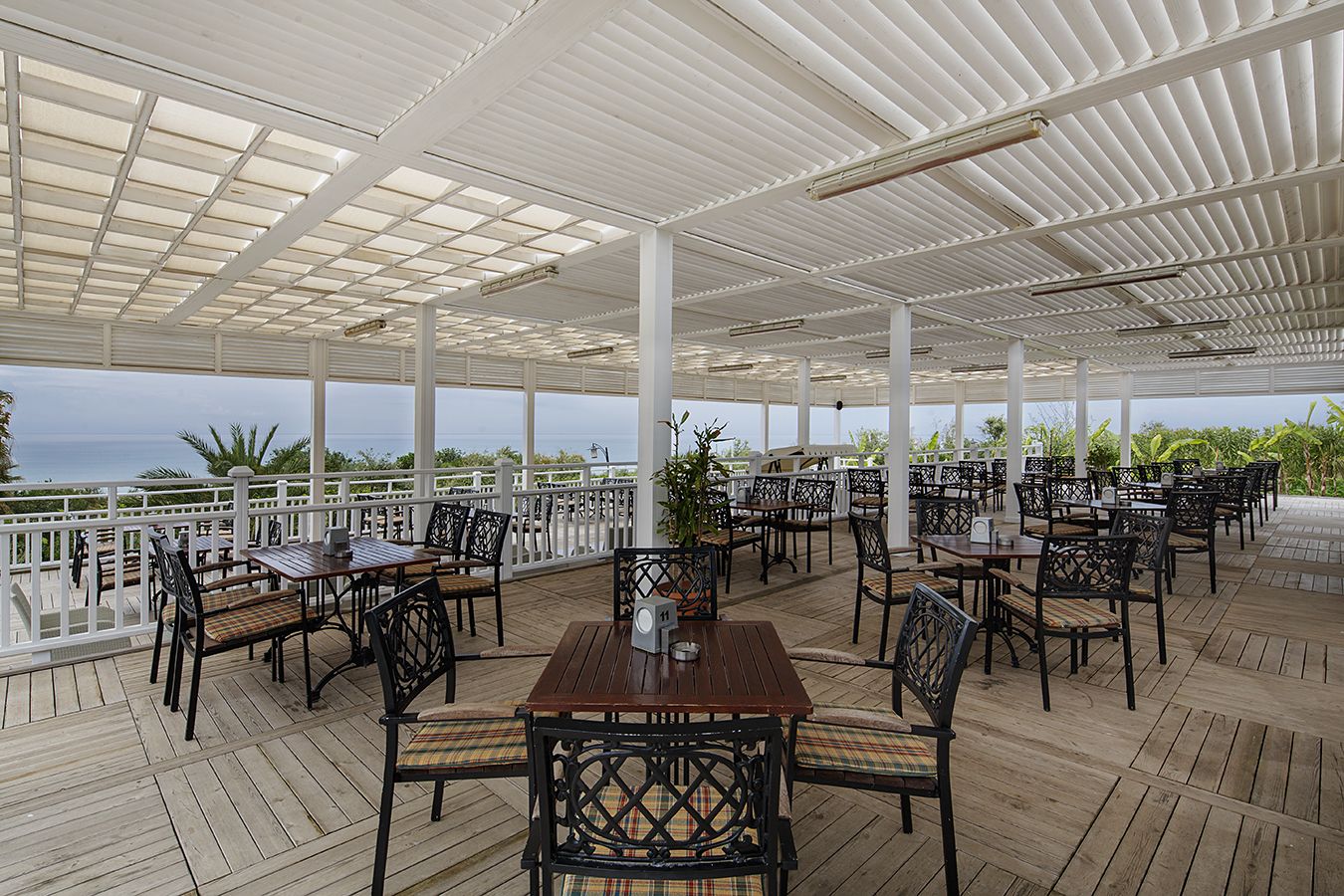 Un patio al aire libre con mesas y sillas de madera, barandal blanco y vista al océano.