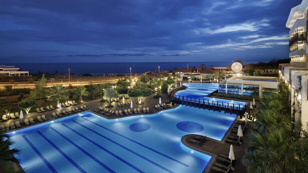 A large, illuminated swimming pool with multiple lanes, surrounded by lounge chairs and umbrellas, set against a backdrop of a clear night sky and a distant view of the ocean. - Alba Royal Hotel - Ultra All Inclusive - Adults Only 16+ (Manavgat)