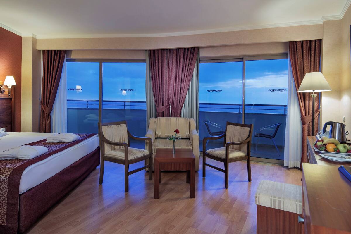 A hotel room with a bed, two armchairs, a small table, a TV, and a balcony with a view of the sea.