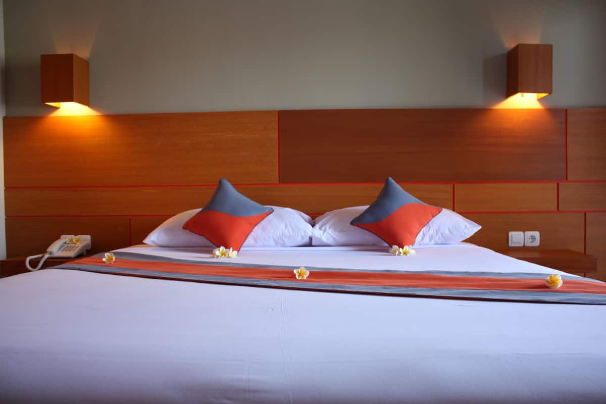 Ein Hotelzimmer mit einem hölzernen Kopfteil, zwei Nachttischlampen, einem Telefon und einem Bett mit weißer Bettwäsche sowie orangefarbenen und grauen Kissen.