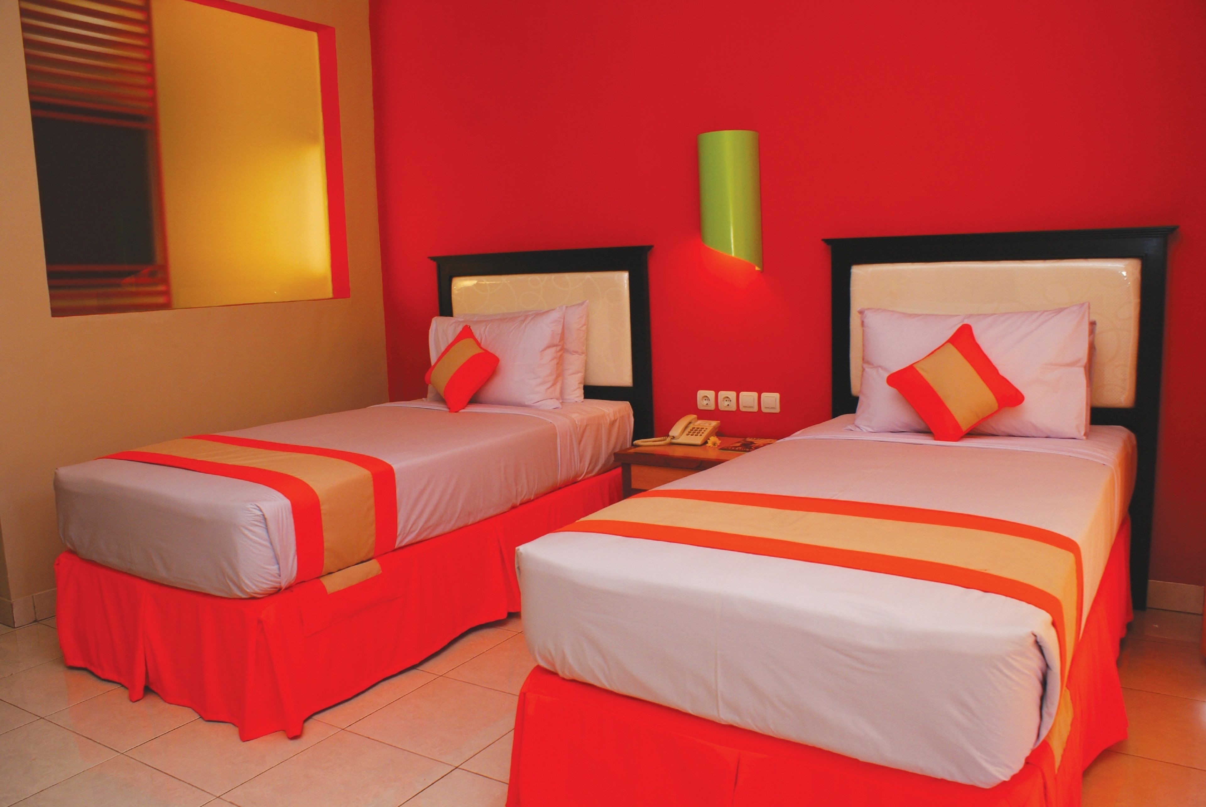 Habitación de hotel con dos camas, cada una con colcha y almohadas rojas y blancas. Hay una lámpara de pared verde sobre las camas y un teléfono en la mesita de noche entre ellas.