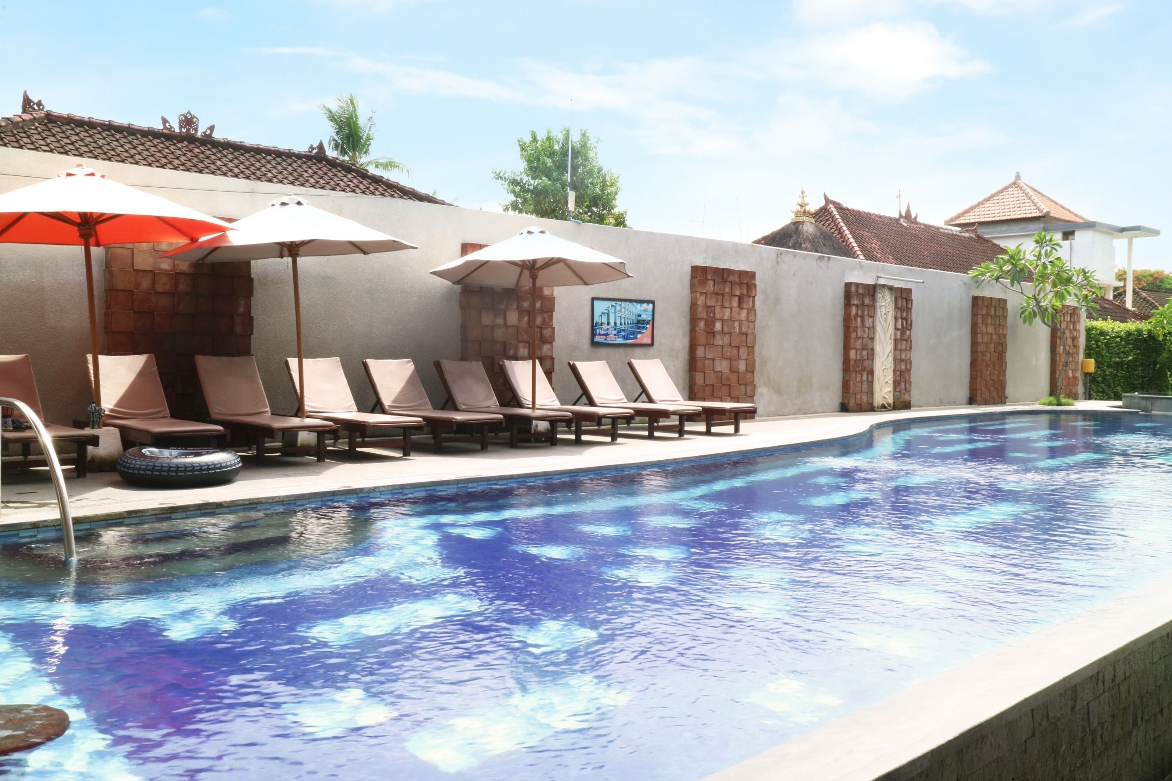 Une piscine avec des chaises longues et des parasols, un toit en tuiles et un mur en briques orné d’un tableau encadré.