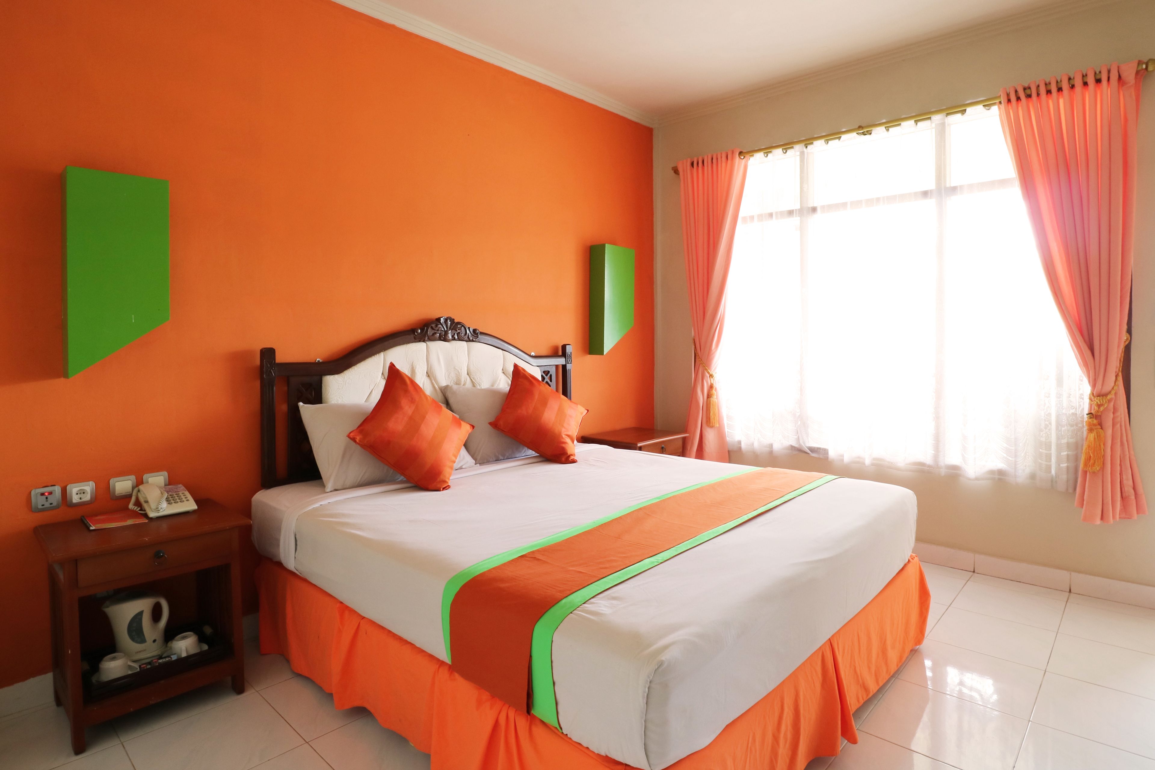 Hotelkamer met een oranje muur, een bed met witte beddengoed en oranje accenten, een nachtkastje met telefoon en waterkoker, en een raam met roze gordijnen.