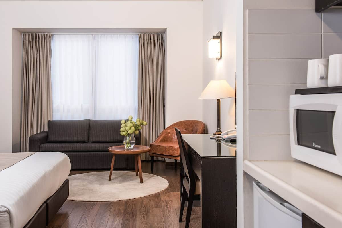A hotel room with a bed, a sofa, a small round table with a vase, a lamp, a chair, and a microwave.