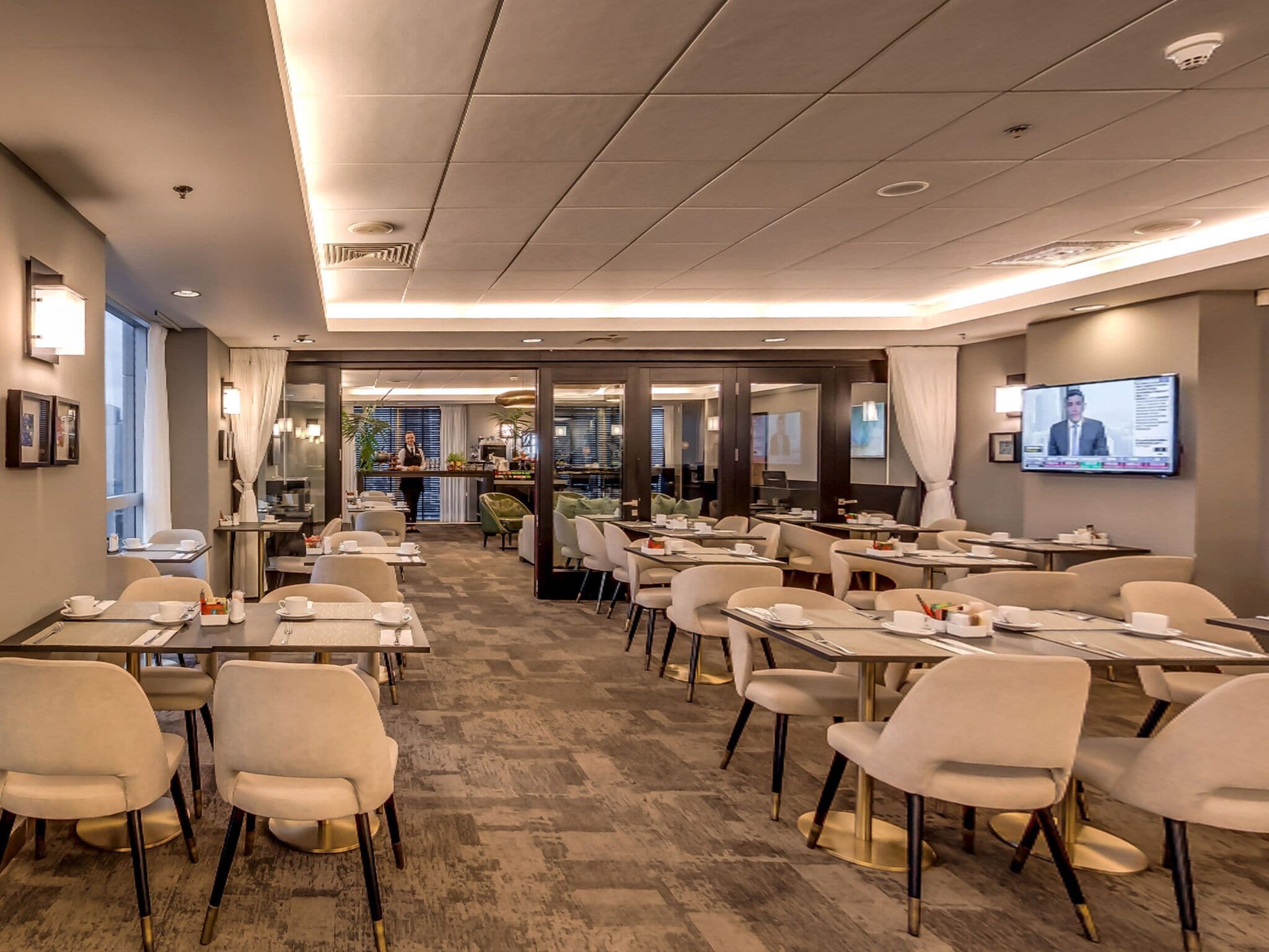 A modern hotel dining area with multiple tables and chairs, a service counter, and a television screen displaying a news program.