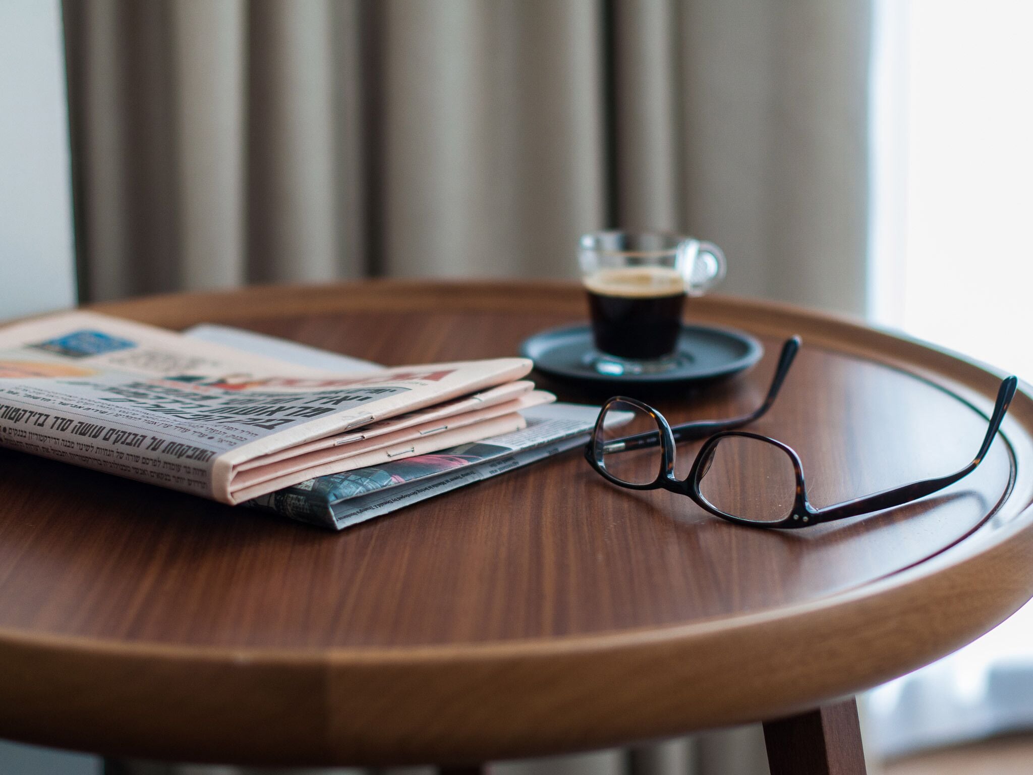 A round wooden table with a newspaper, a pair of glasses, and a cup of coffee.