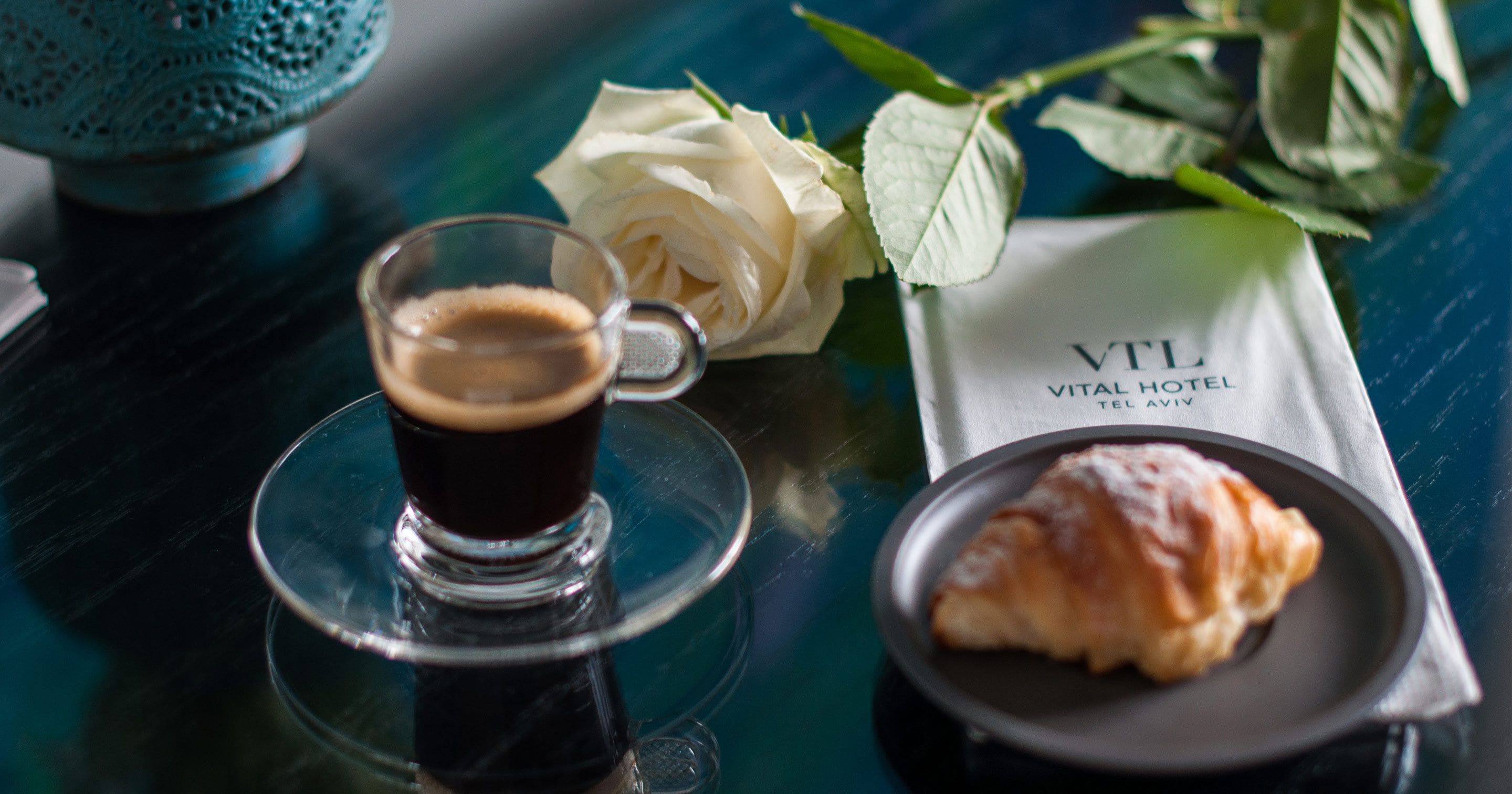 A glass of coffee, a croissant, a white rose, and a napkin with 'VITAL HOTEL' text on a table.