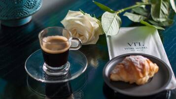 A glass of coffee, a croissant, a white rose, and a napkin with 'VITAL HOTEL' text on a table.