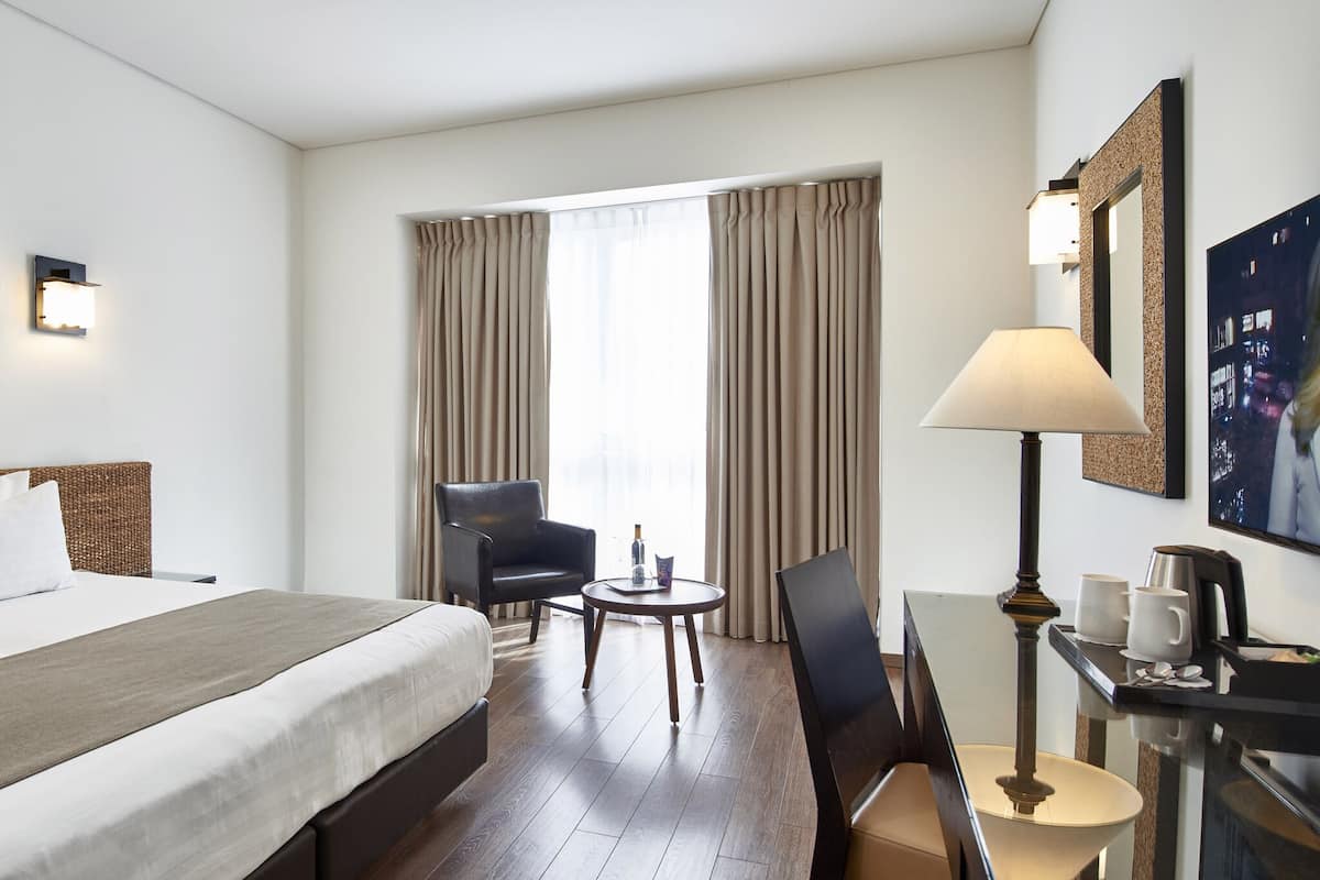 Une chambre d’hôtel avec un lit, un bureau avec une lampe, une chaise, une petite table et une fenêtre avec des rideaux.