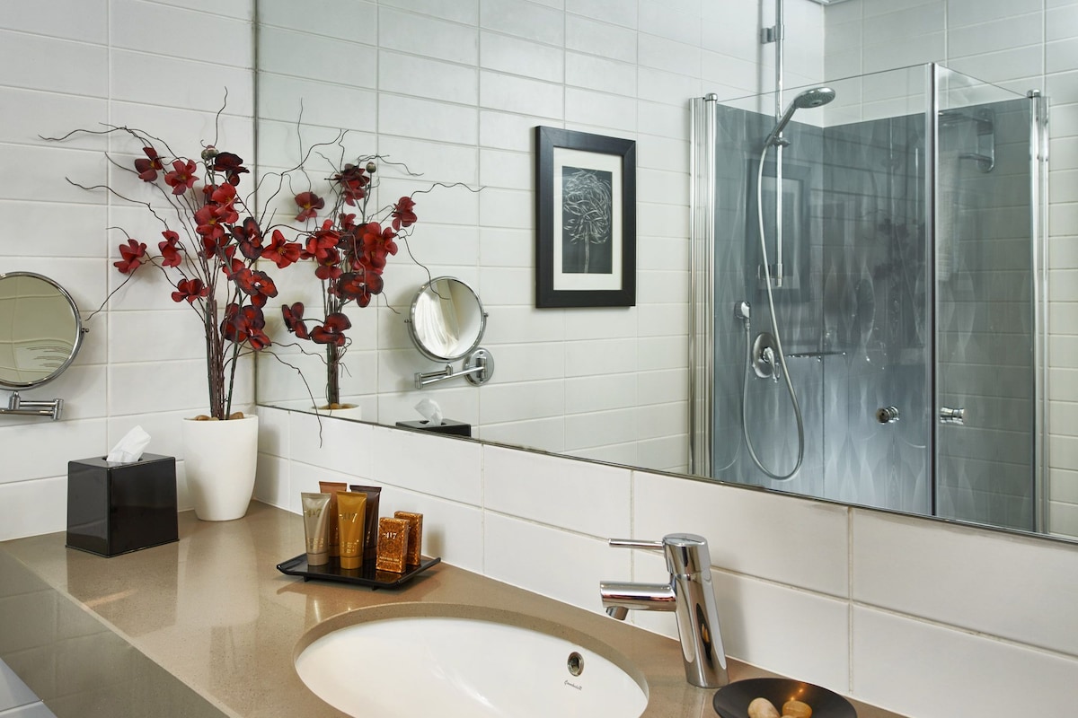 A bathroom with a white sink, a glass shower enclosure, a mirror, and a vase with red flowers.