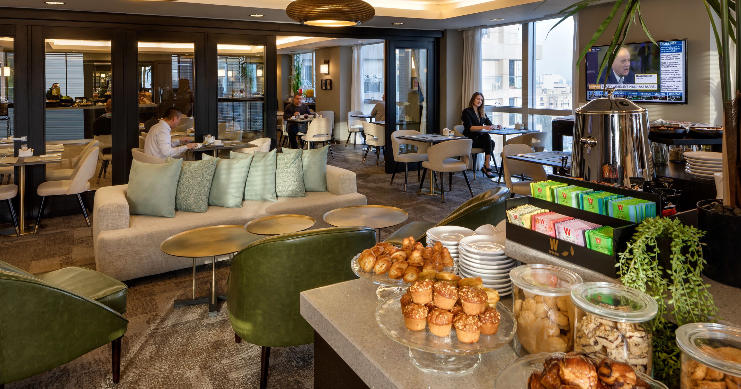 Un hall de hotel con zona de estar, bufé de desayuno y un televisor que muestra noticias.