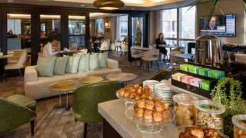 Un hall de hotel con zona de estar, bufé de desayuno y un televisor que muestra noticias.