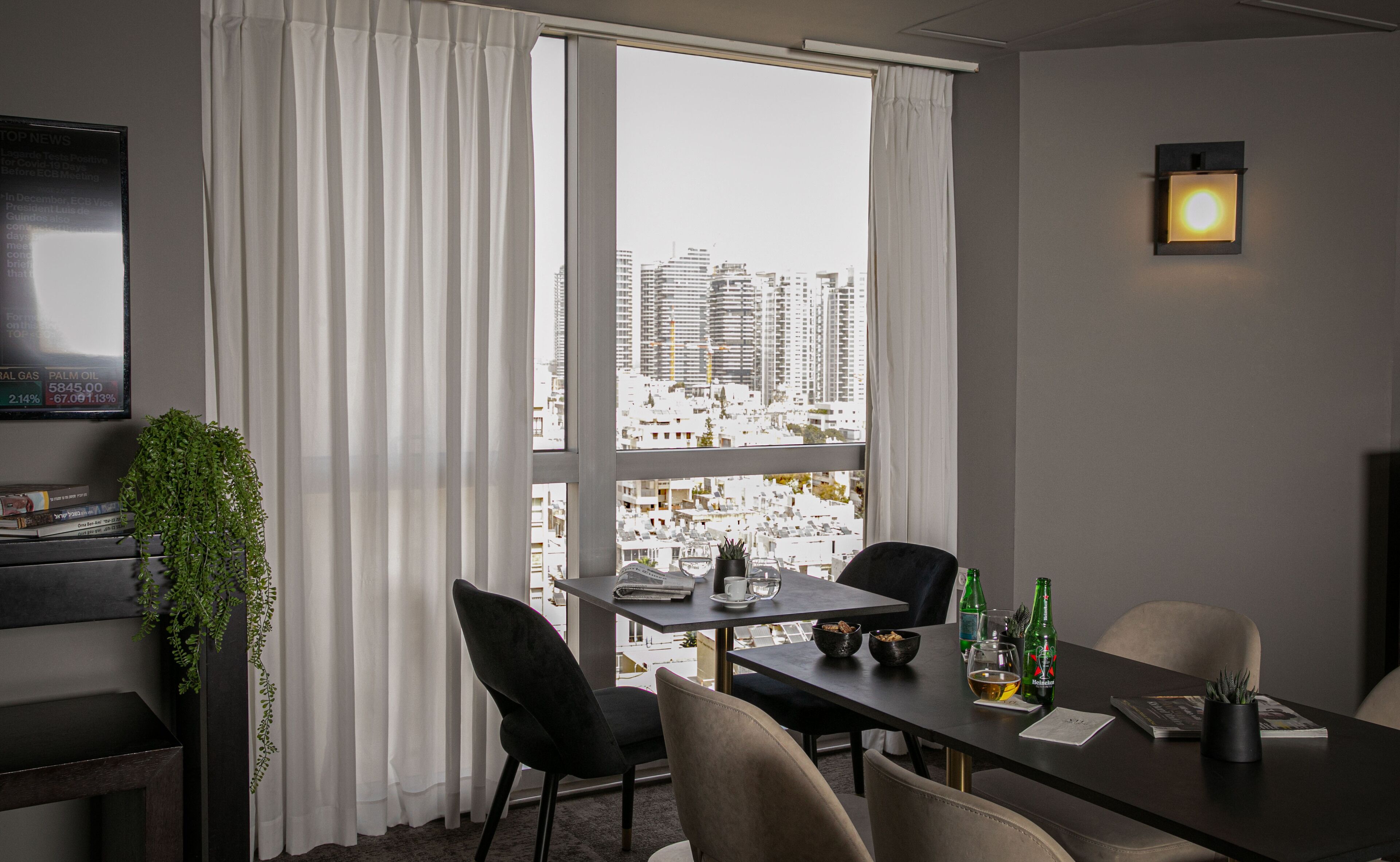 Une chambre d’hôtel moderne dotée d’une grande fenêtre donnant sur la ville, d’un coin repas avec une table dressée pour deux personnes et d’une petite plante posée sur la table d’appoint.