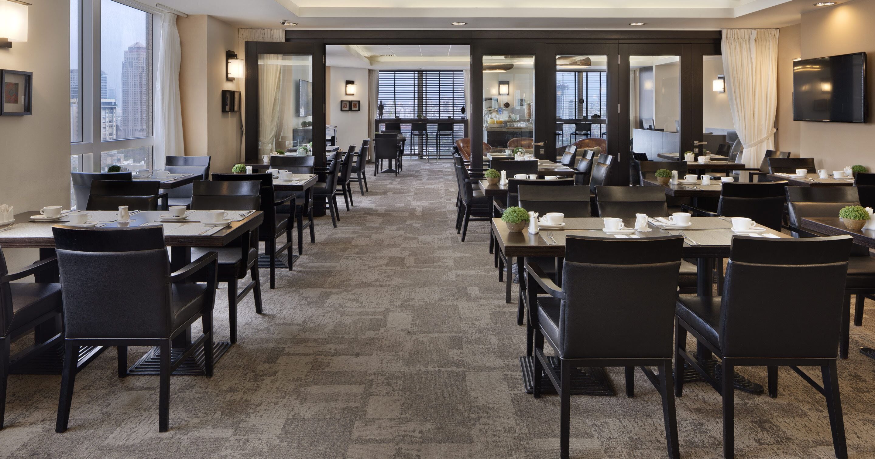 Un comedor de hotel con varias mesas y sillas, ventanas amplias y vista a la ciudad.