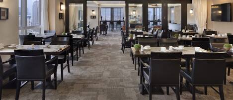 Un comedor de hotel con varias mesas y sillas, ventanas amplias y vista a la ciudad.