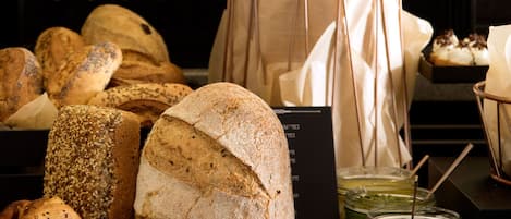 A variety of breads and pastries displayed on a wooden table, with a jar of jam and a menu stand in the background.