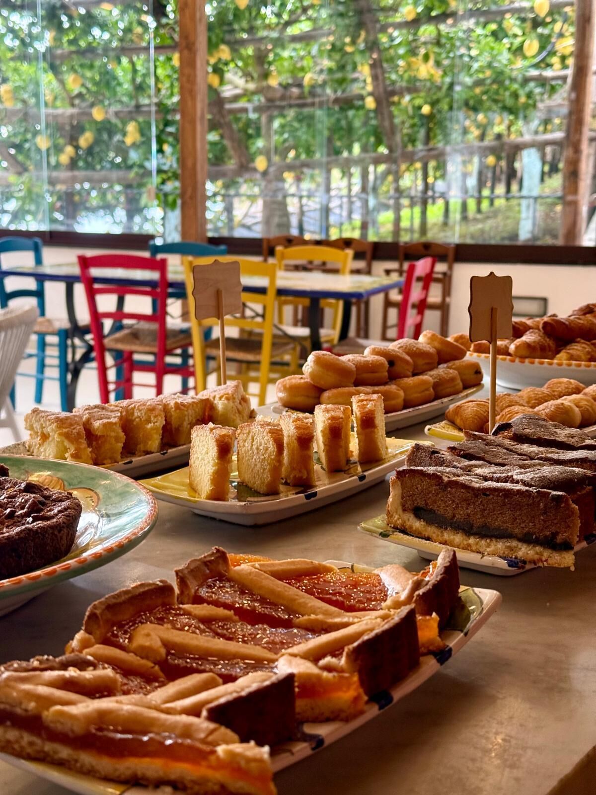 Une variété de desserts est disposée sur une table dans une pièce lumineuse, agrémentée de chaises colorées et offrant une vue sur la verdure à l’extérieur.