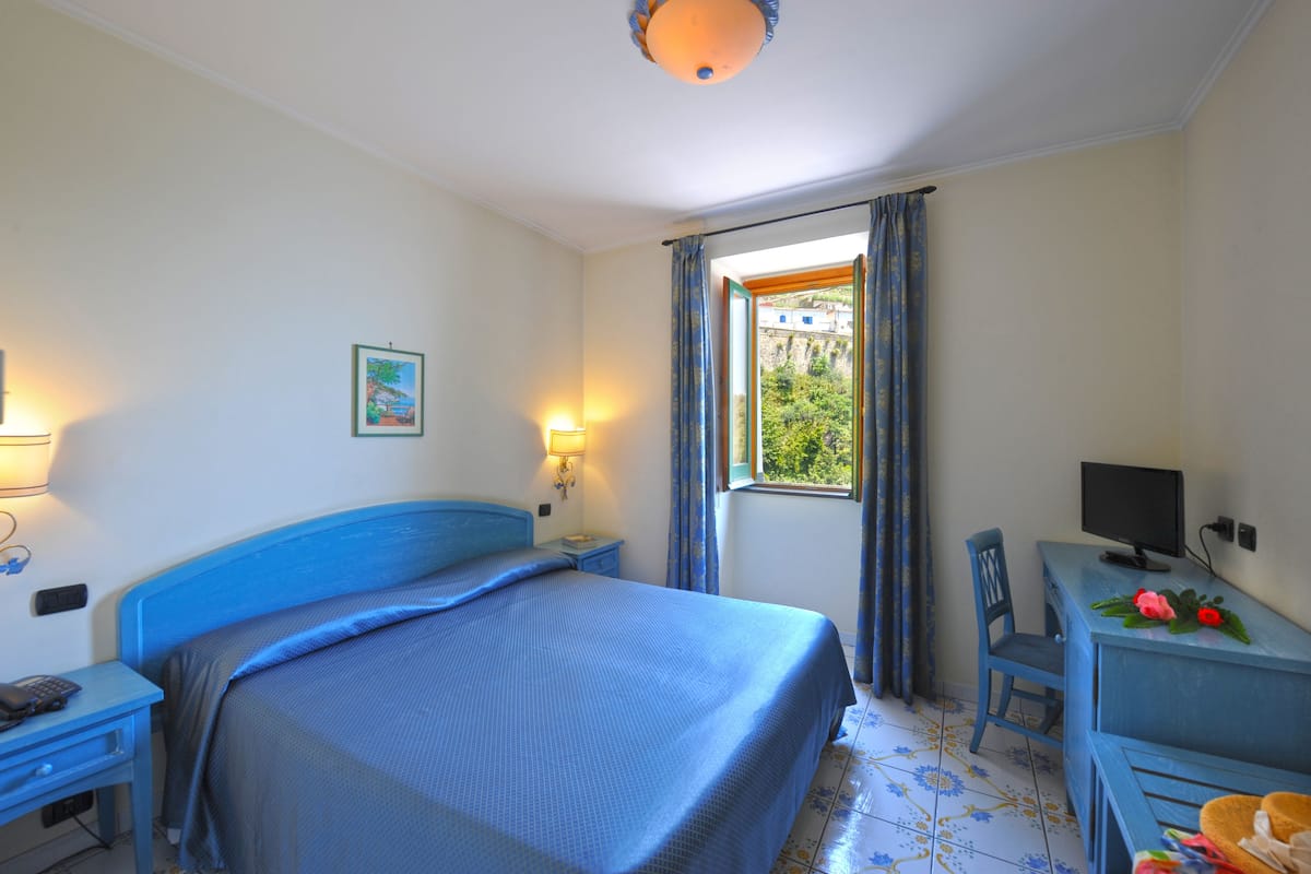 Une chambre à coucher comprenant un lit, un bureau, une chaise, un téléviseur et une fenêtre avec des rideaux.