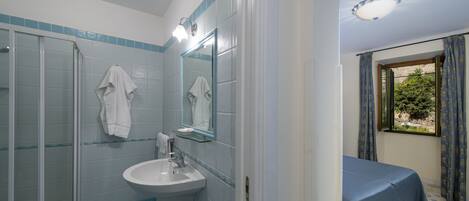 Un baño con cabina de ducha de vidrio, un lavabo y un espejo. En la habitación contigua, hay una cama con cubrecama azul.
