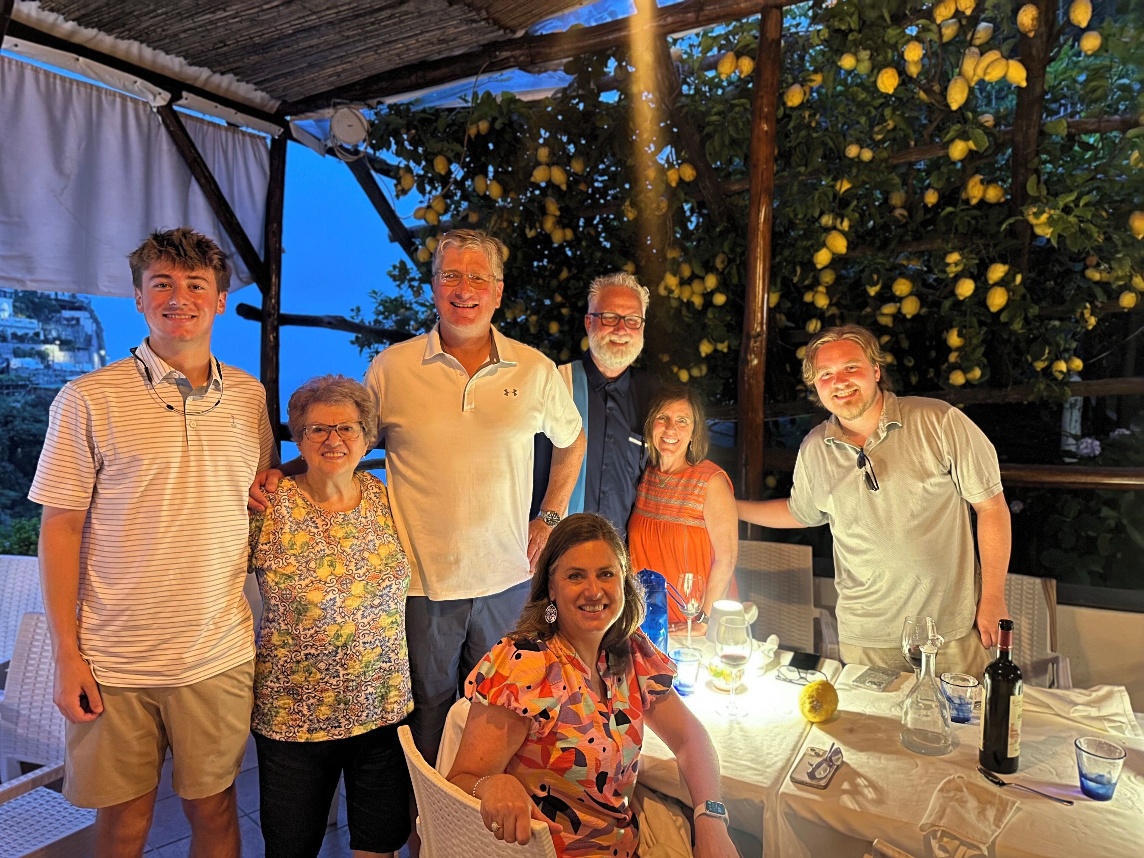 A group of people gathered around a table with a bottle of wine, glasses, and a lemon on a tablecloth.