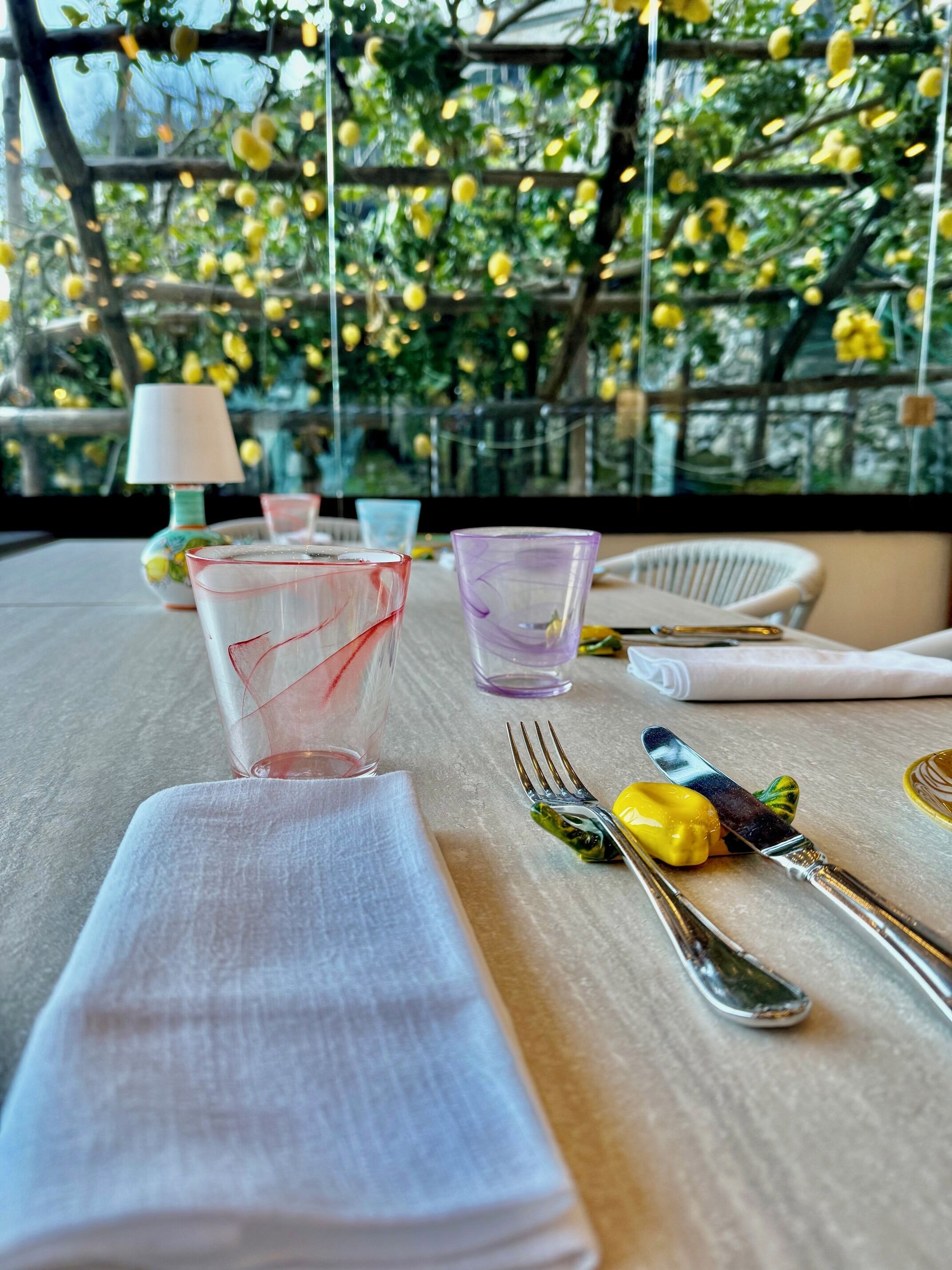 A table set with clear glasses, a fork, and a knife, with a lamp and a chair in the background.