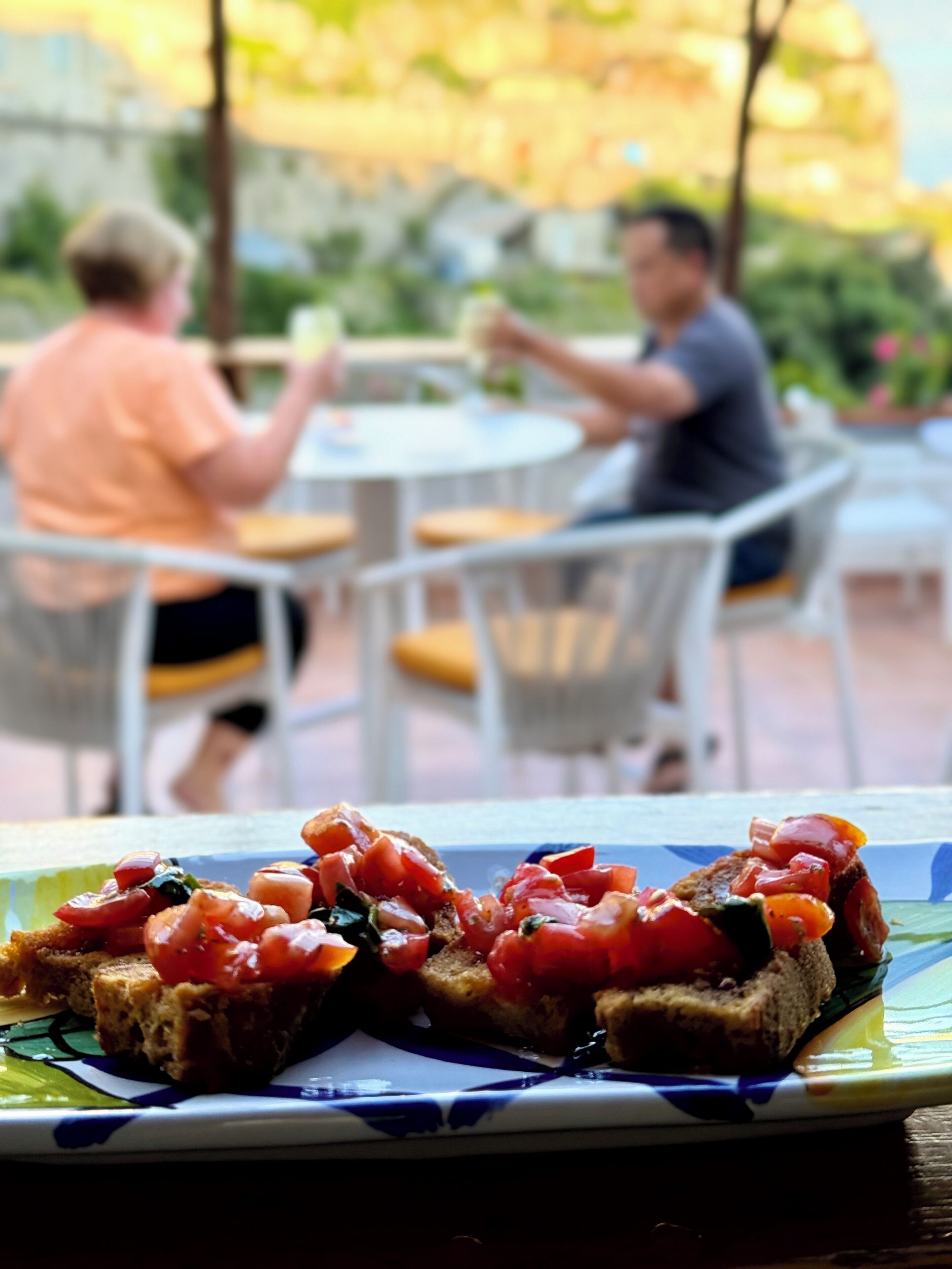 A dining area with outdoor tables and chairs, a plate of bruschetta with tomatoes and basil, and two people seated at a table.