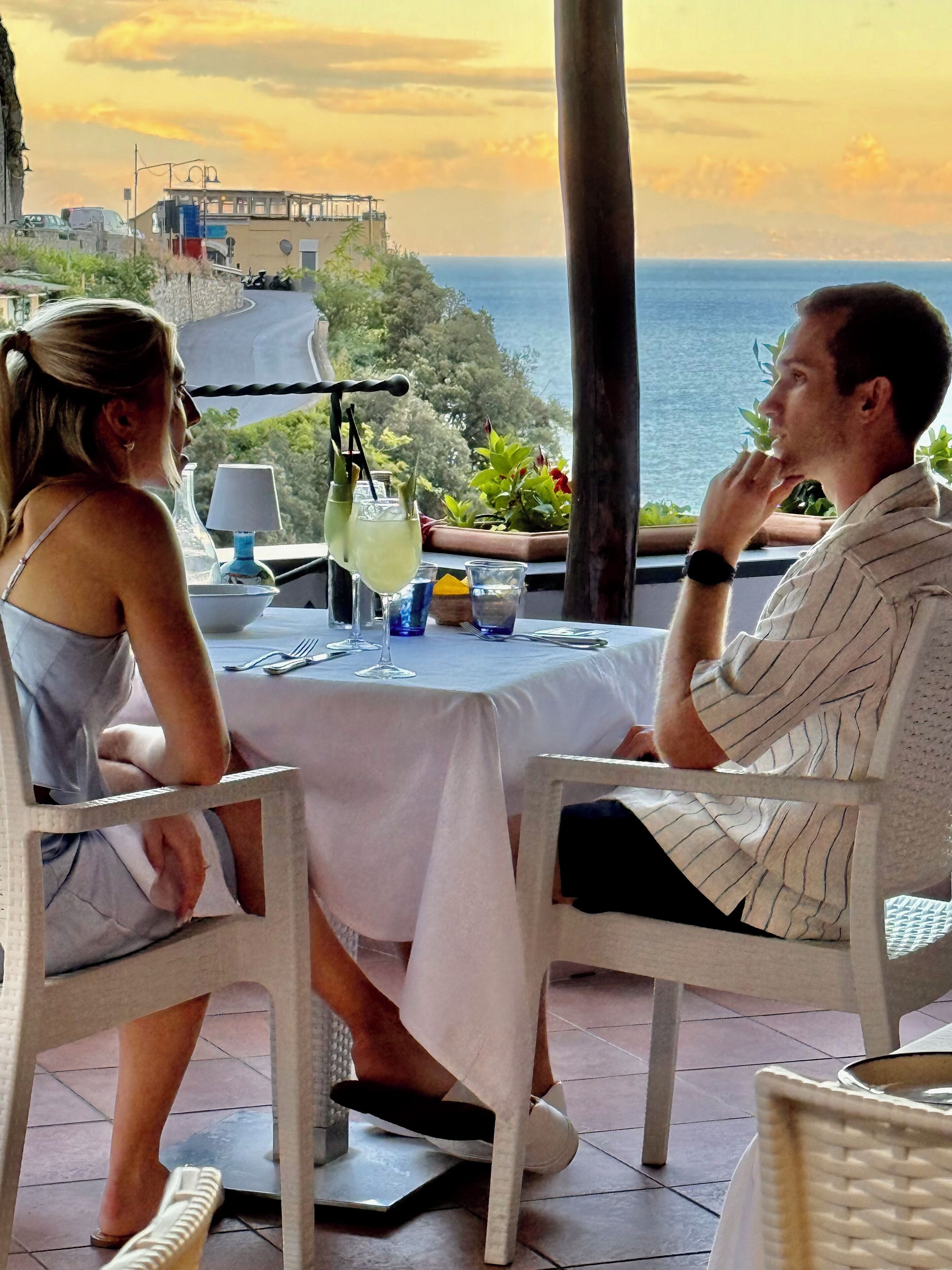 A couple dining at a table with a view of the sea and a building in the background.