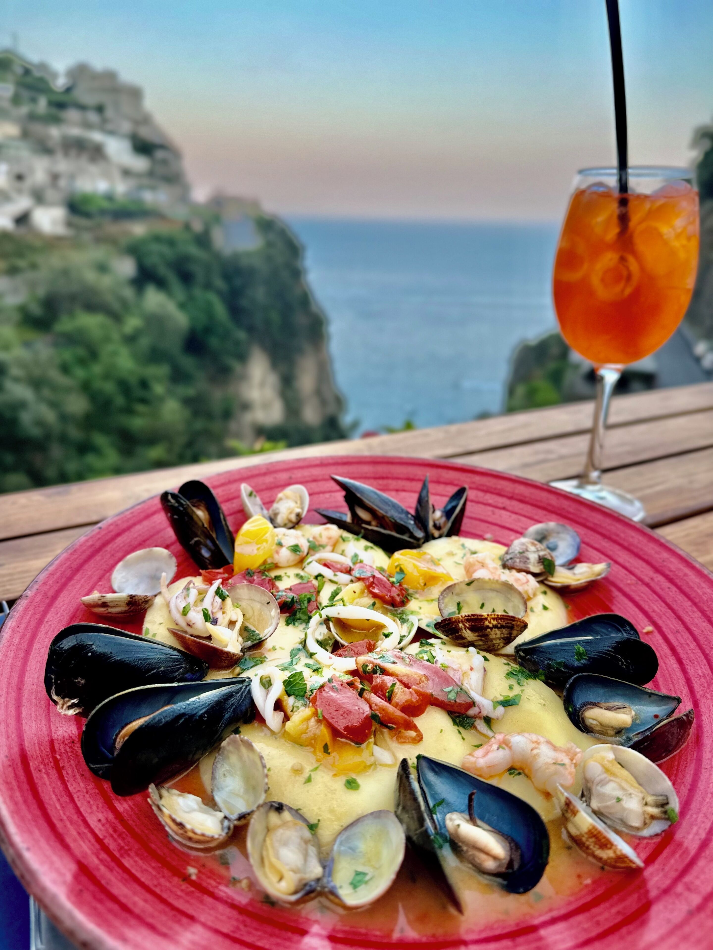 Plato de mariscos sobre un fondo de acantilados con vista panorámica, acompañado de un vaso de bebida anaranjada.