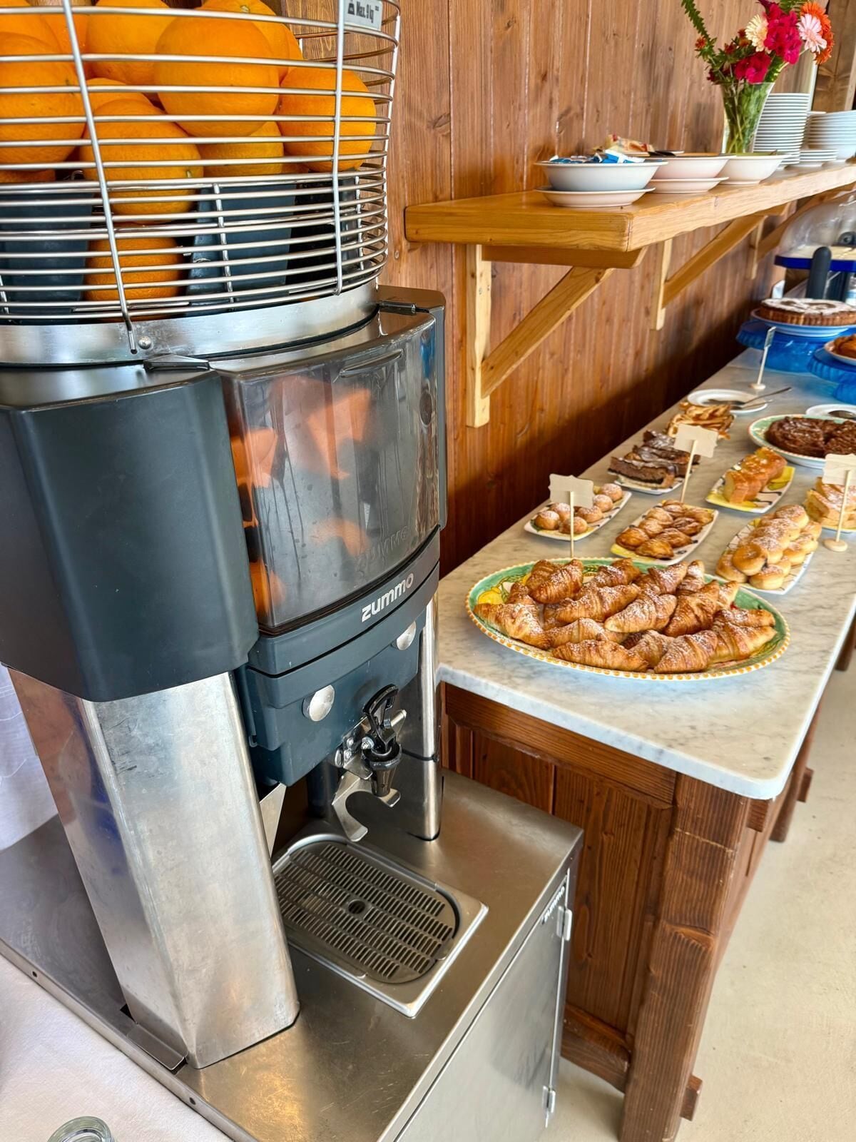 Un coin cuisine équipé d’une machine à expresso de la marque Zummo, d’un panier de fruits et d’une table de buffet garnie de viennoiseries.