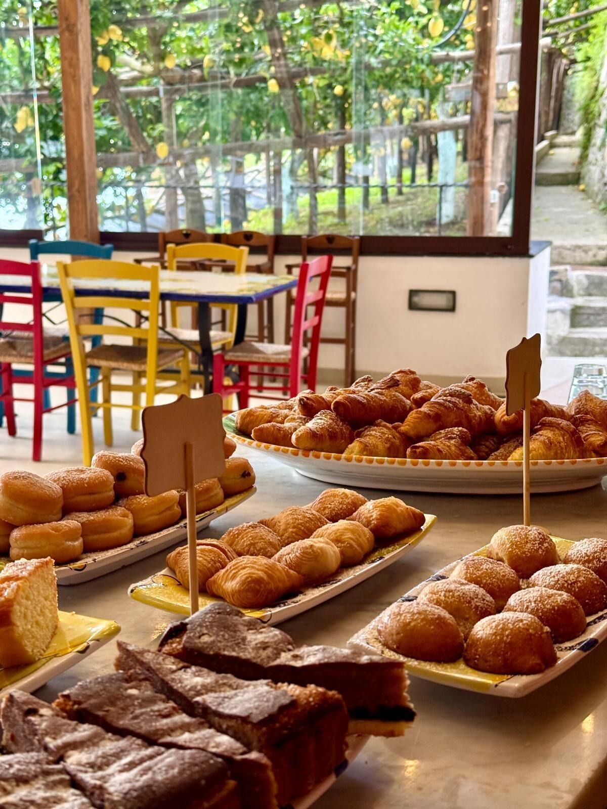Un bufé de desayuno con pasteles y pan, sillas coloridas y vistas a zonas verdes a través de amplias ventanas.