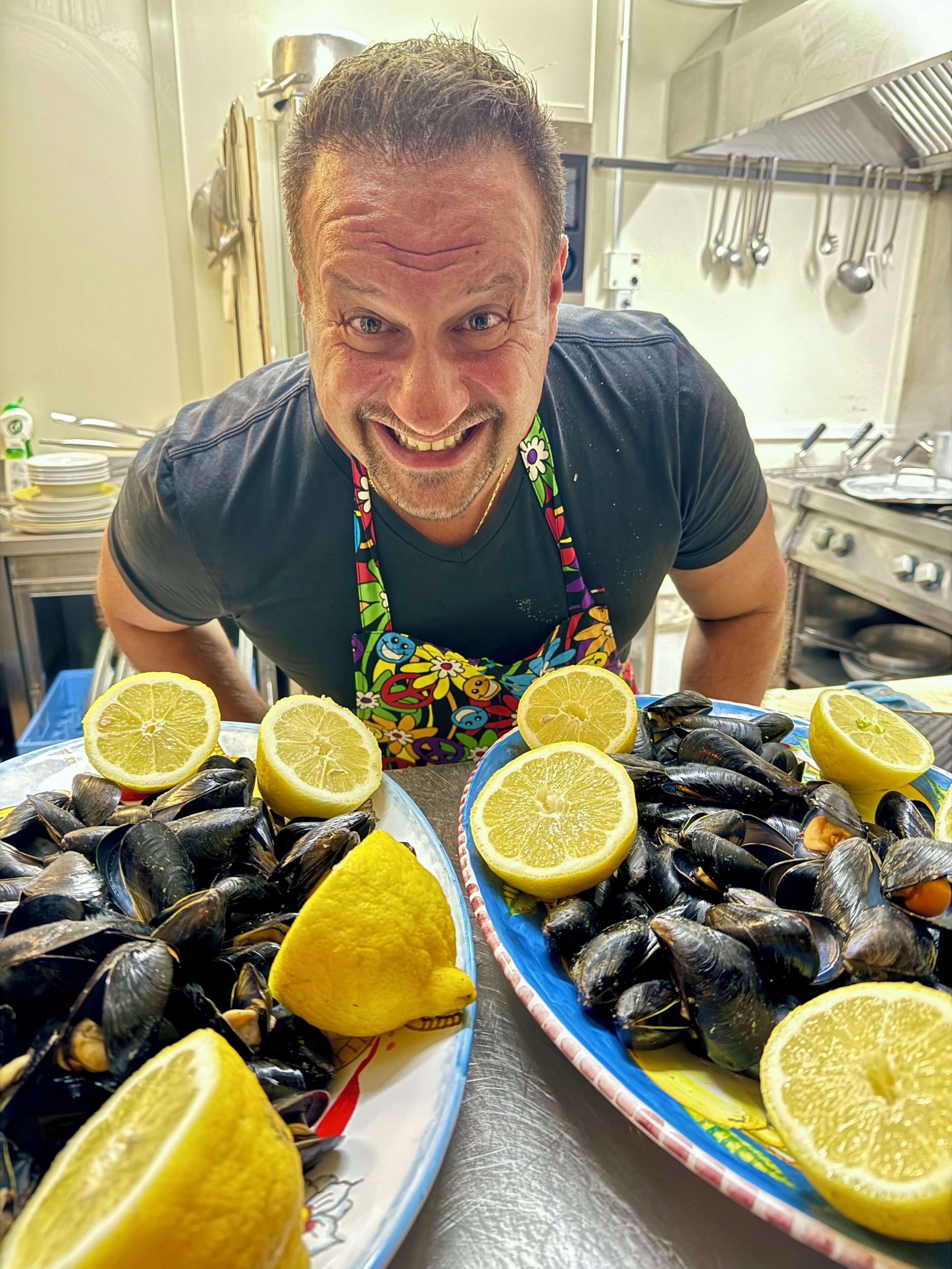A person in a kitchen with two large plates of mussels and lemon slices.