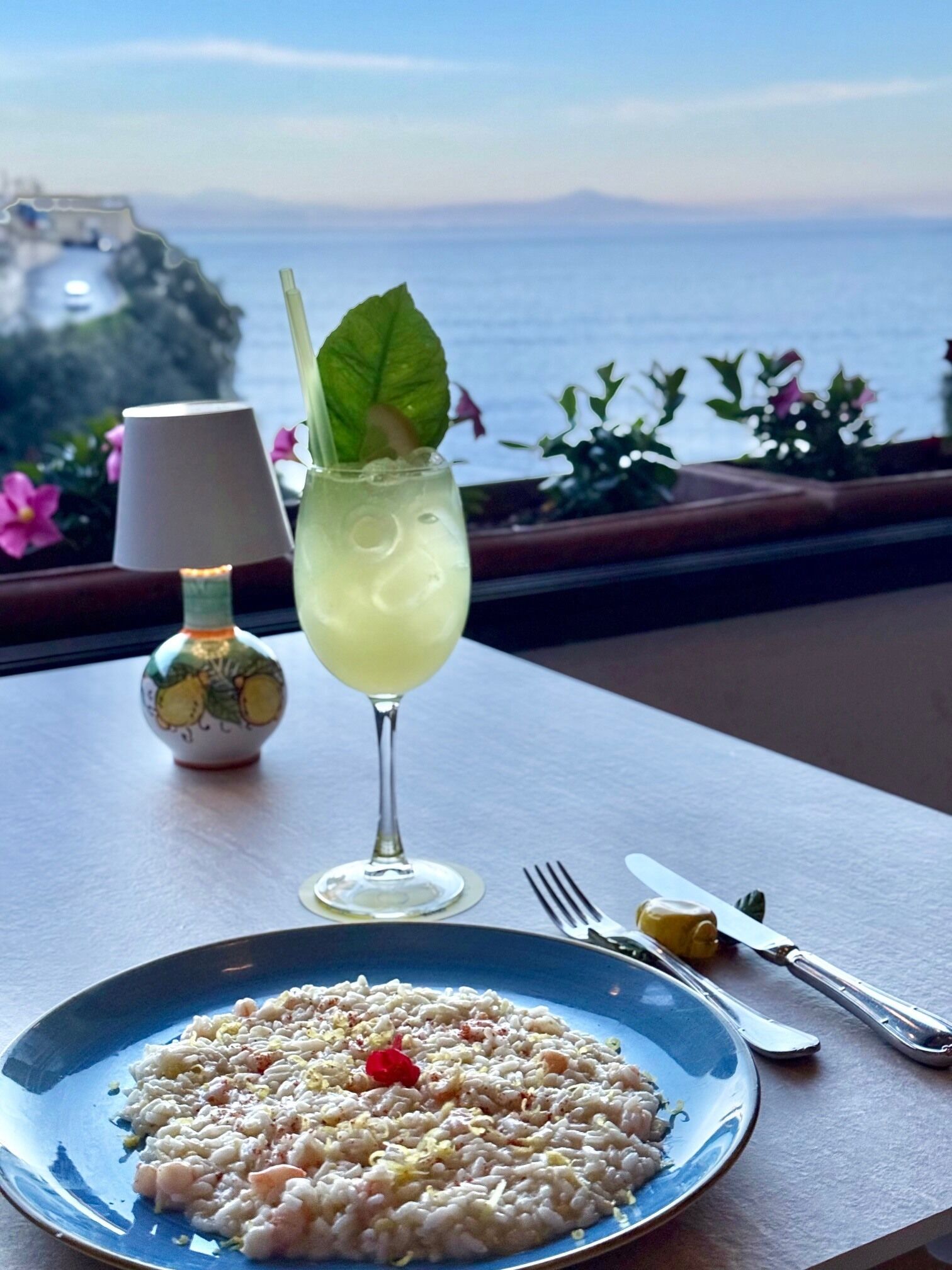 A table with a plate of risotto, a cocktail with a mint leaf, and a lamp.