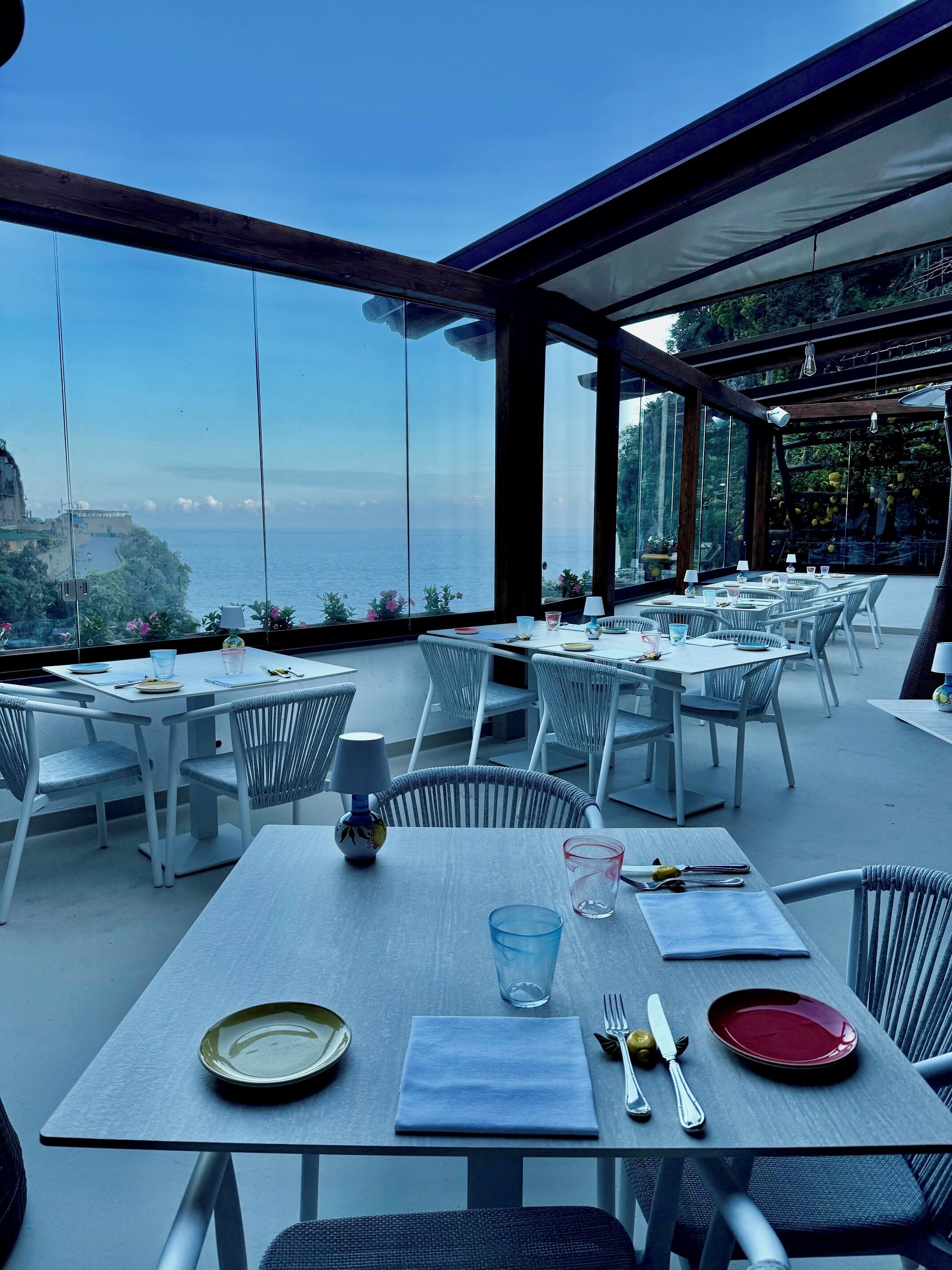 An outdoor dining area with tables and chairs, overlooking a scenic view of the sea and a distant coastline.