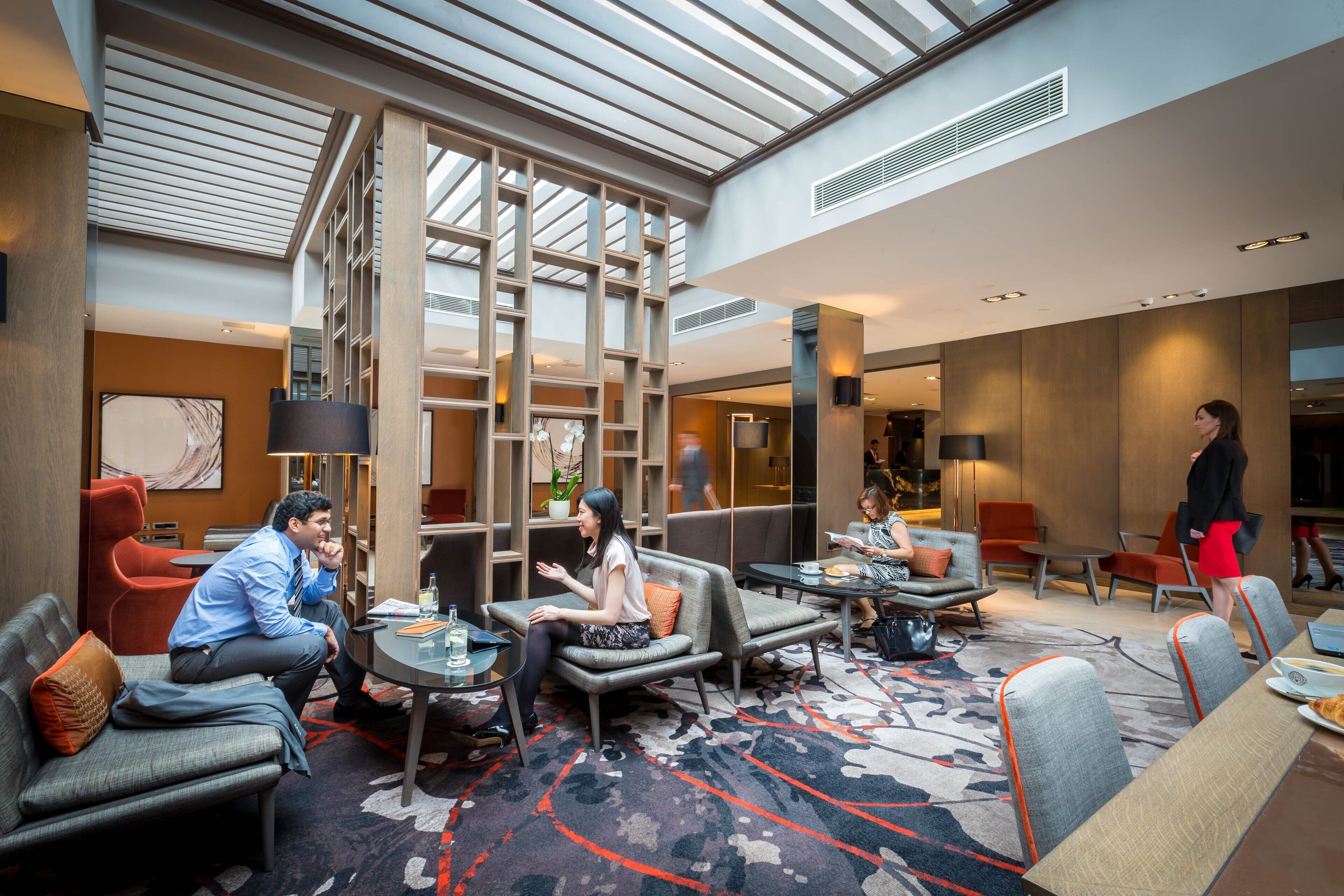 A modern hotel lobby with a high ceiling, a central seating area with sofas and tables, and a large window providing natural light.