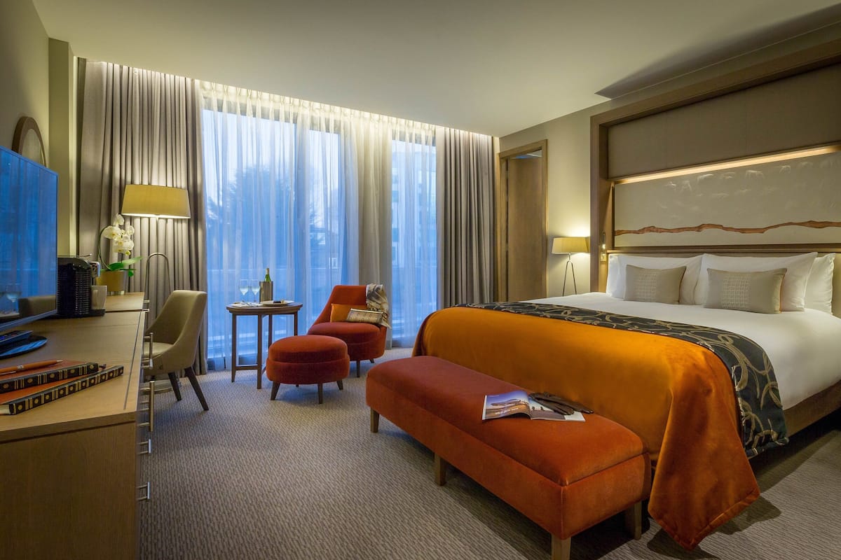 A hotel room with a large bed, a desk with a computer, a chair, a small table with a lamp, and a window with curtains.