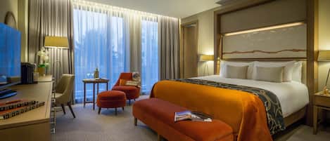 A hotel room with a large bed, a desk with a computer, a chair, a small table with a lamp, and a window with curtains.