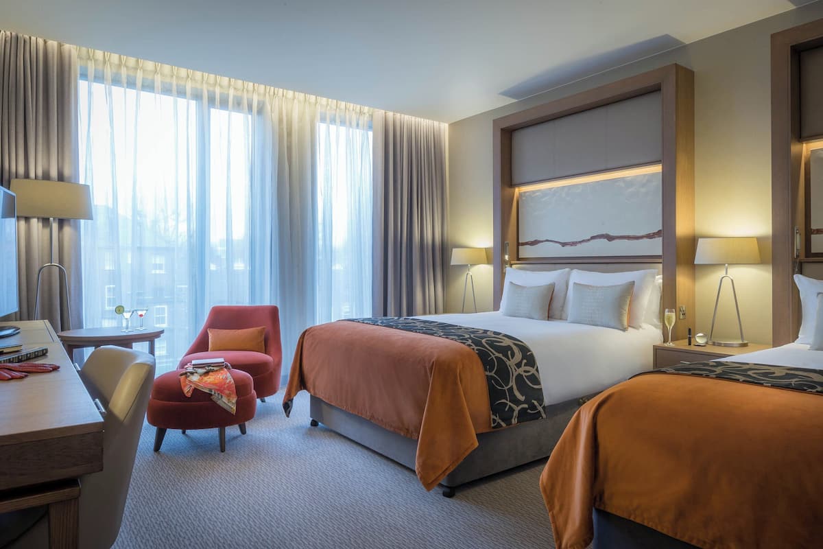 A hotel room with a large bed, two bedside tables, a desk, a chair, and a view of the city through large windows.