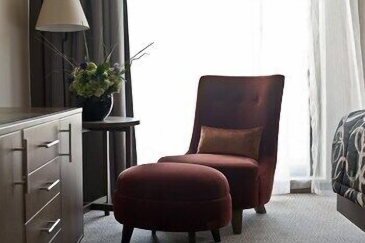 A hotel room with a dark-colored armchair, a matching ottoman, a side table with a lamp, and a potted plant.