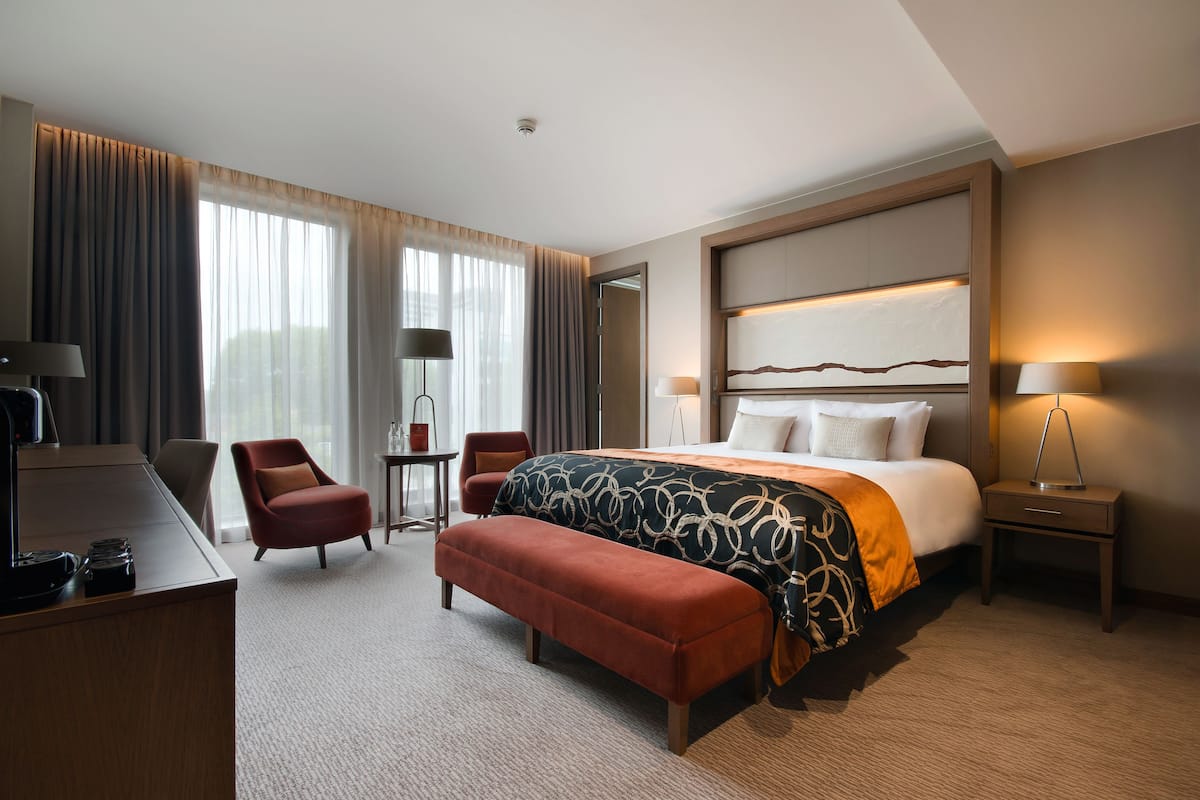 A hotel room with a large bed, a desk, a chair, a small table, and a bench.