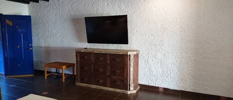 Una habitación con un televisor de pantalla plana montado en una pared blanca texturizada, un mueble de madera con cajones, un taburete pequeño de madera y una puerta azul.