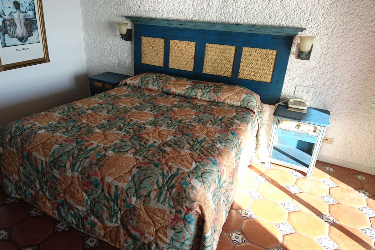 A bedroom with a floral bedspread, a blue headboard with woven panels, a nightstand with a phone, and a wall-mounted light fixture.