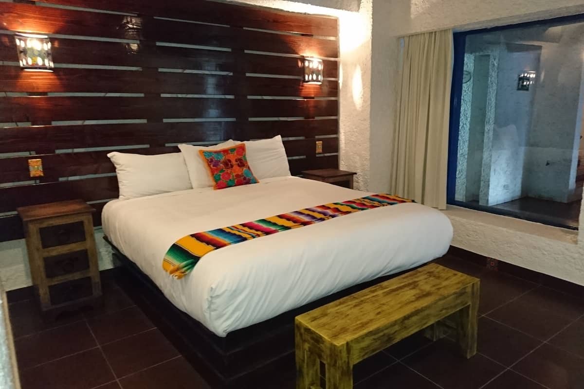 A hotel room with a wooden headboard, a large bed with white linens, a colorful throw blanket, a wooden bench, and a window with a view of the outside.