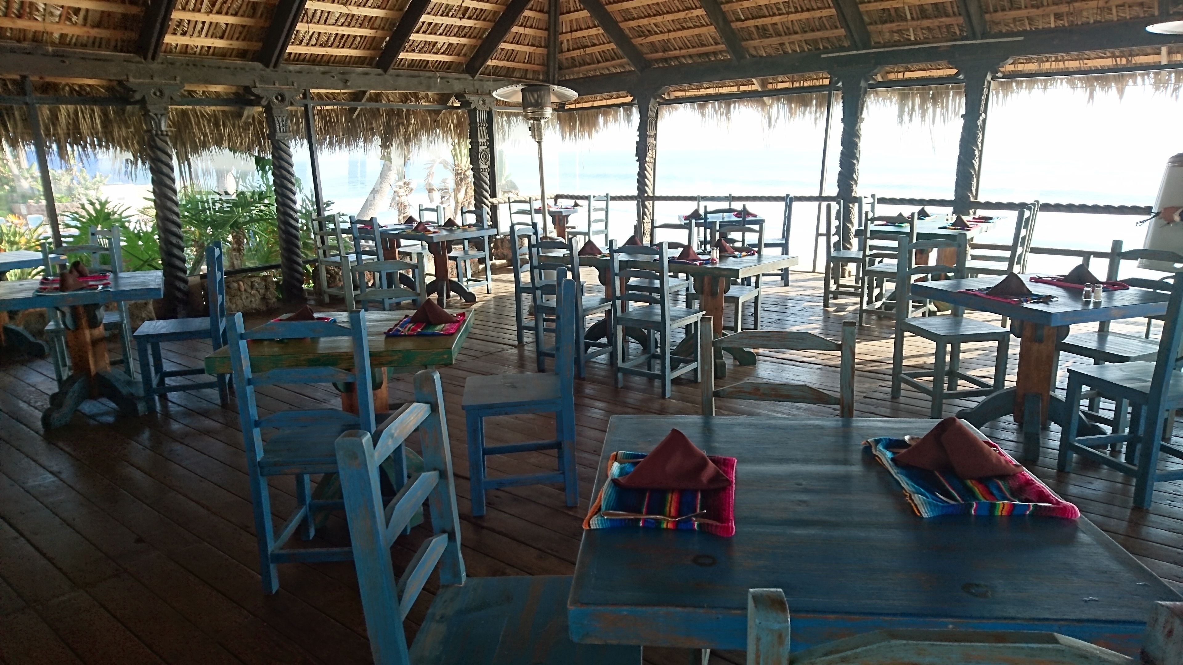 A covered outdoor dining area with wooden tables and chairs, some with red napkins.