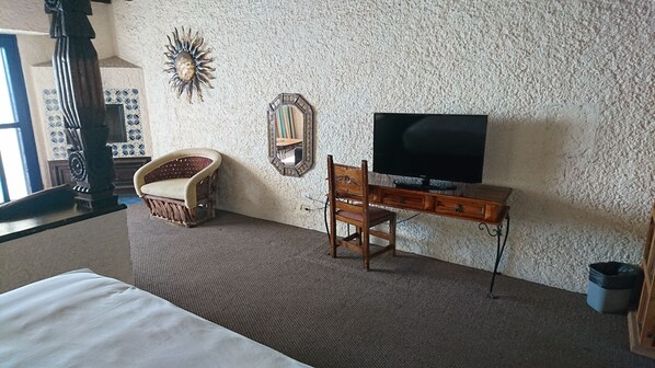 Un dormitorio con una cama, un escritorio de madera con televisor, una silla, un espejo decorativo y un adorno de pared con forma de sol.
