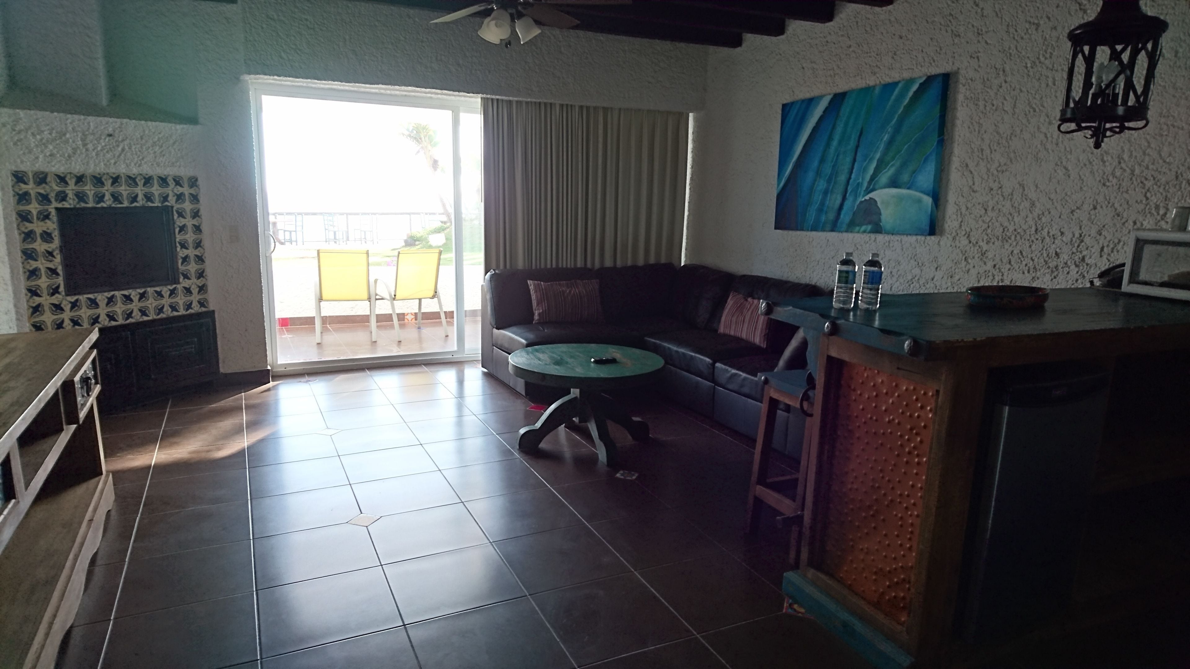A living room with a sofa, a coffee table, and a TV. There is a sliding glass door leading to an outdoor area with chairs.