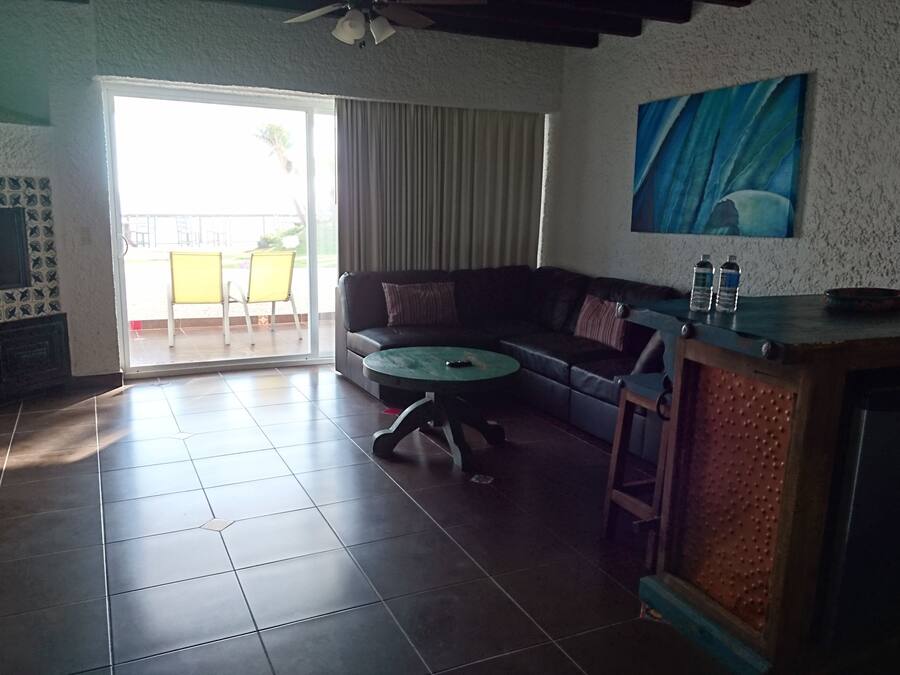 A living room with a sofa, a coffee table, and a TV. There is a sliding glass door leading to an outdoor area with chairs.