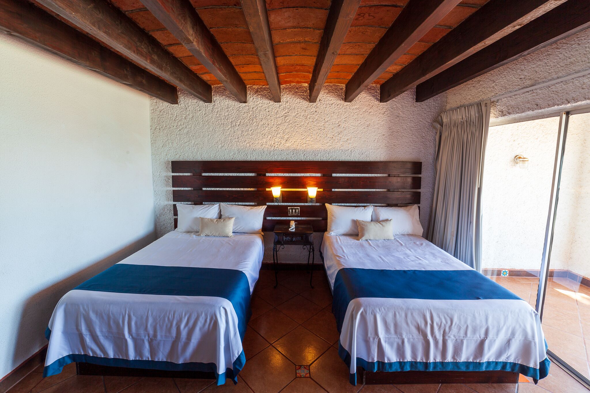 A room with two beds, a wooden ceiling, a nightstand, and a window with curtains.