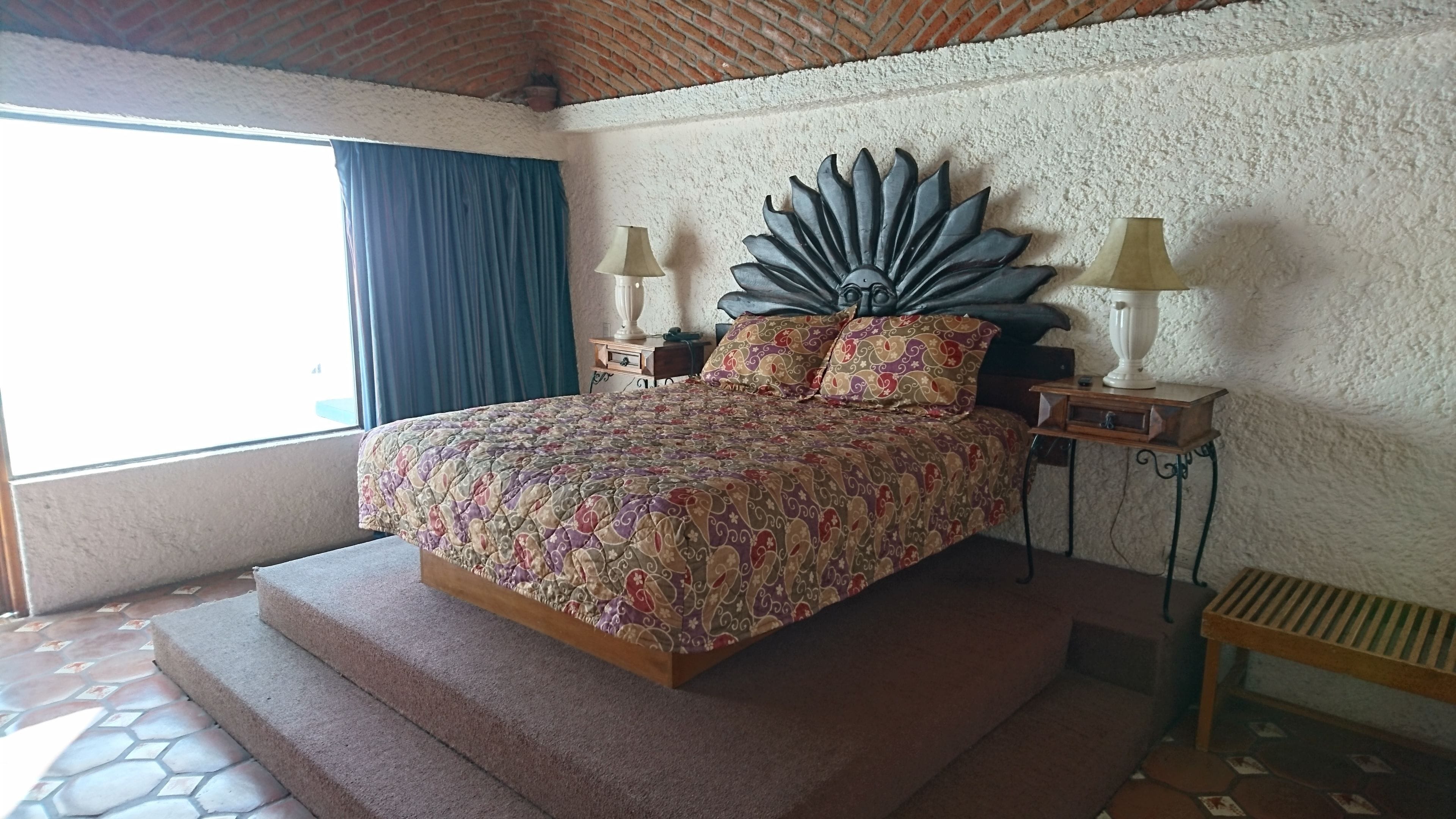A bedroom with a large bed, a decorative wall piece, two bedside tables with lamps, and a view of the outdoors through a window with blue curtains.