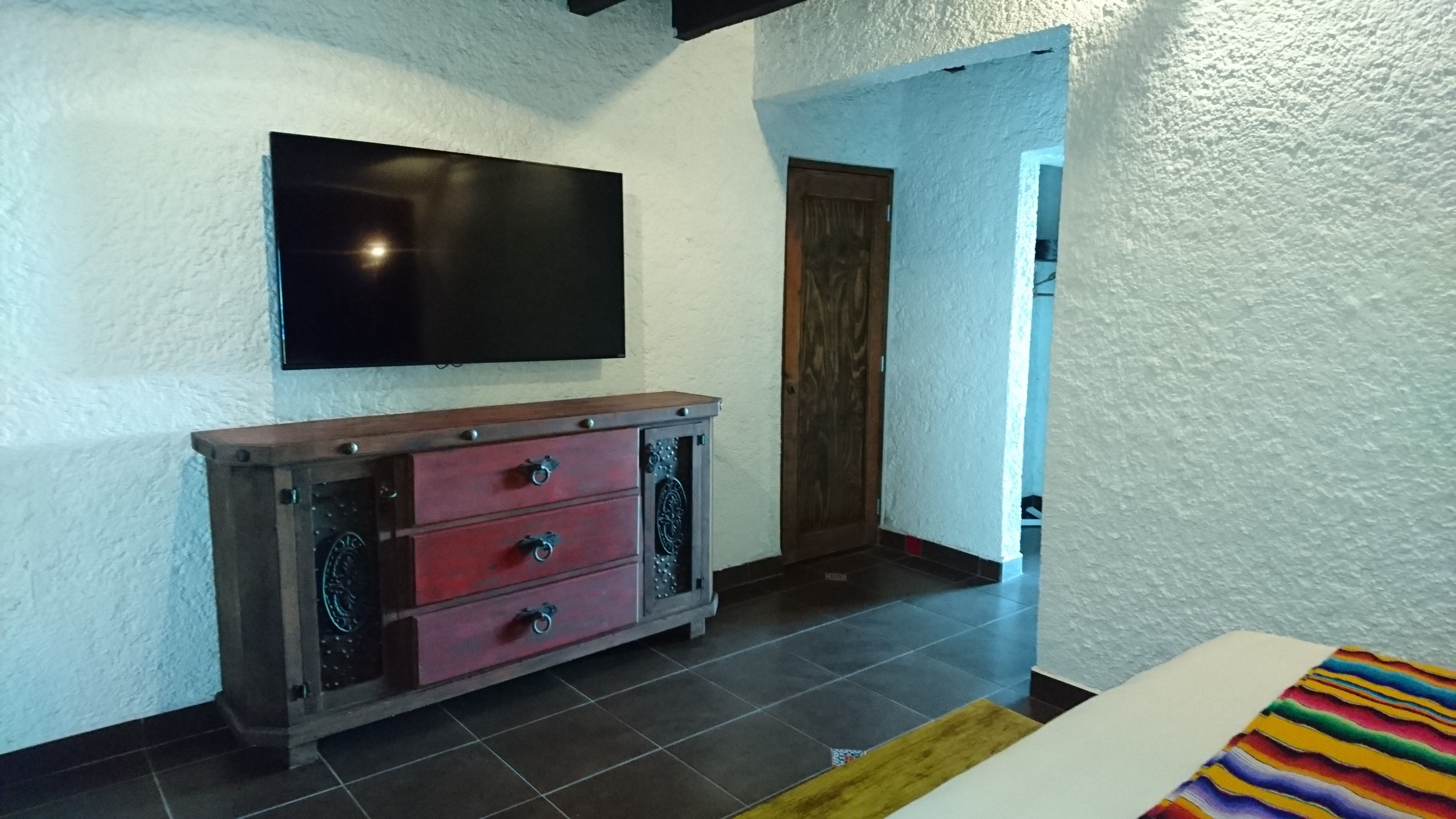 A room with a flat-screen TV mounted on the wall, a wooden cabinet with drawers, and a door leading to another room.
