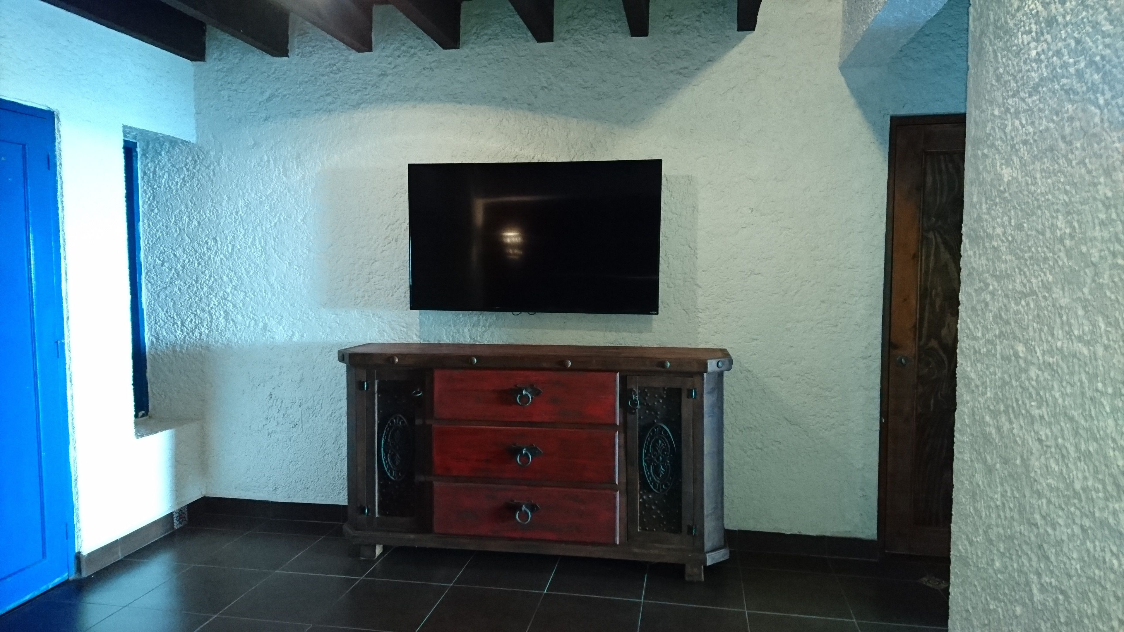 A room with a flat-screen TV mounted on a textured wall, a wooden cabinet with drawers, and a blue door.